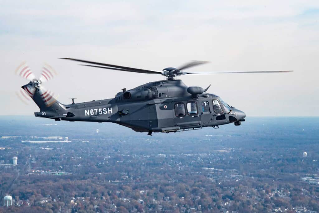 Militärischer Hubschrauber im Flug über eine Landschaft mit Wald und Stadt im Hintergrund.